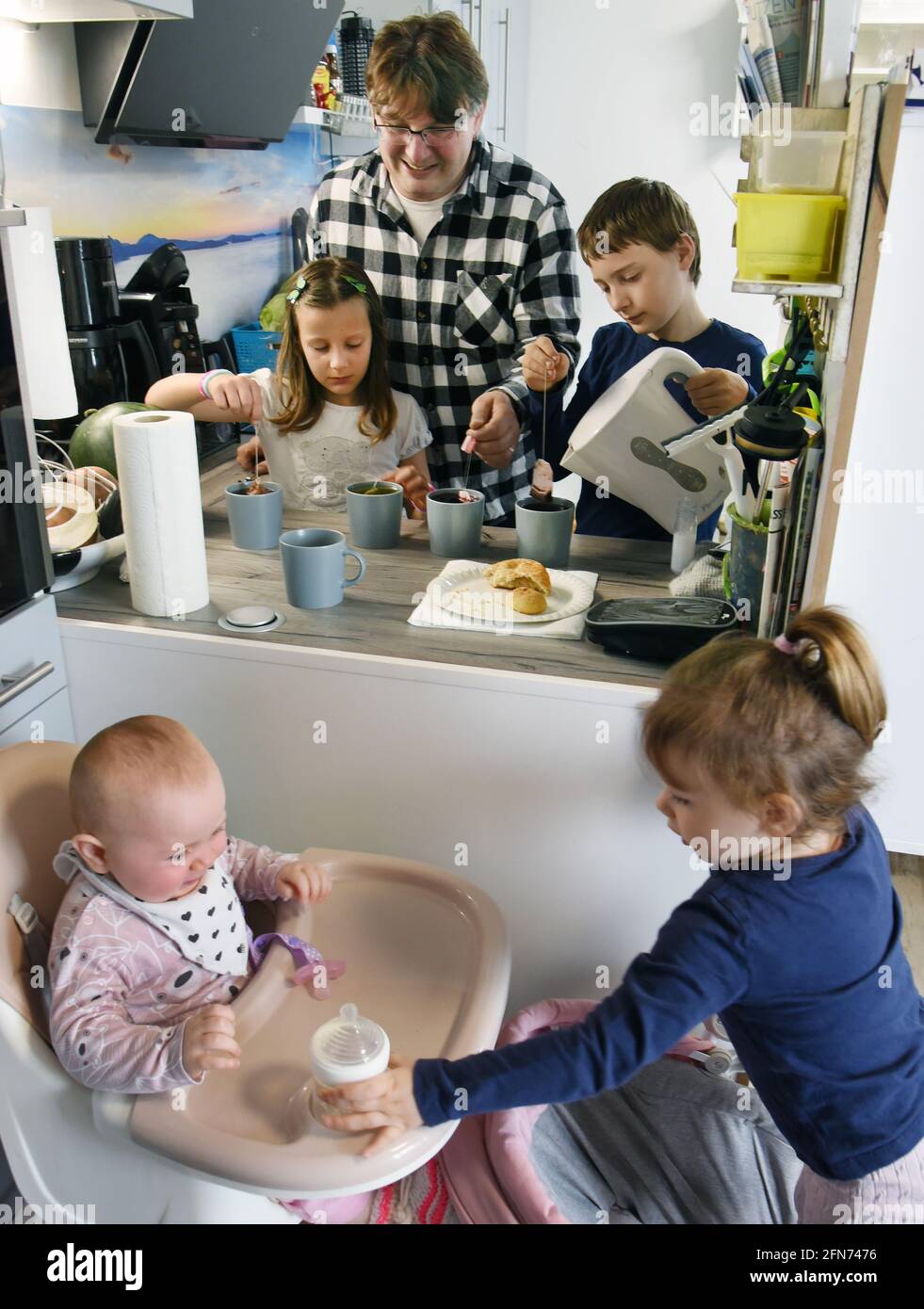 14. Mai 2021, Sachsen, Leipzig: Ronny Steglich bereitet mit den Kindern Timon (r, 11) und Kiana (l, 7) in der Wohnküche ihrer neuen 5-Zimmer-Wohnung Tee zu. Im Vordergrund spielt die 2-jährige Johanna (r) mit ihrer Schwester Paulina (9 Monate). Ronny Steglich, gelernter Koch, Restaurantfachmann und Büroverwalter, und seine Frau Sandra sind Eltern von neun Kindern und konnten vor wenigen Wochen mit sechs ihrer Kinder in ein neu erbautes Mehrfamilienhaus ziehen. Da die drei großen Kinder ihre eigenen Wohnungen in unmittelbarer Nähe haben, feiert die Großfamilie alle Bi Stockfoto