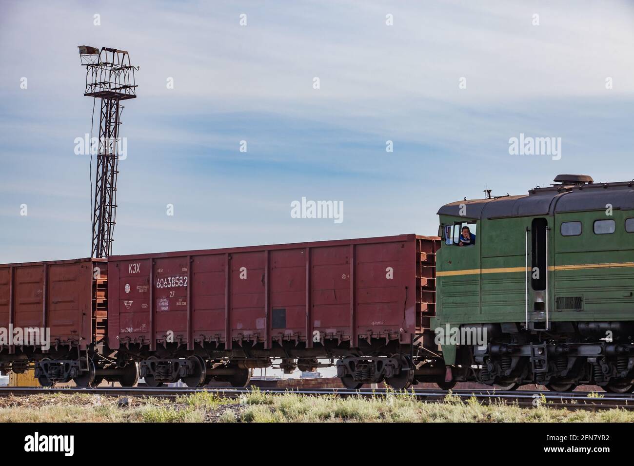 Khromtau, Kasachstan: Erzladestation. Offene Waggons und Lokomotive mit Maschinist (Betreiber) rechts. Lichtmast links. Blauer Himmel, Wolken. Stockfoto