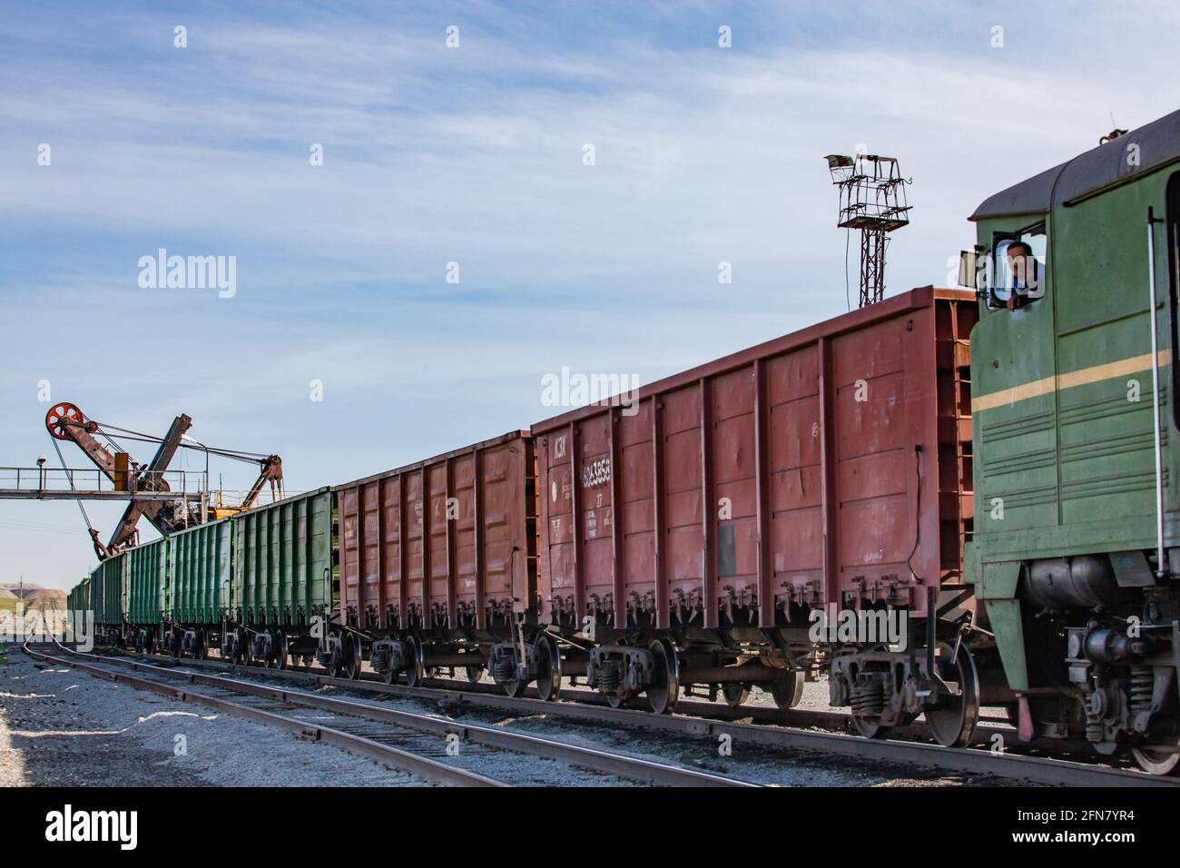 Khromtau, Kasachstan: Erzladestation. Offene Waggons und Bagger. Lokomotive mit lächelndem asiatischen Fahrer rechts. Blauer Himmel, Wolken. Stockfoto