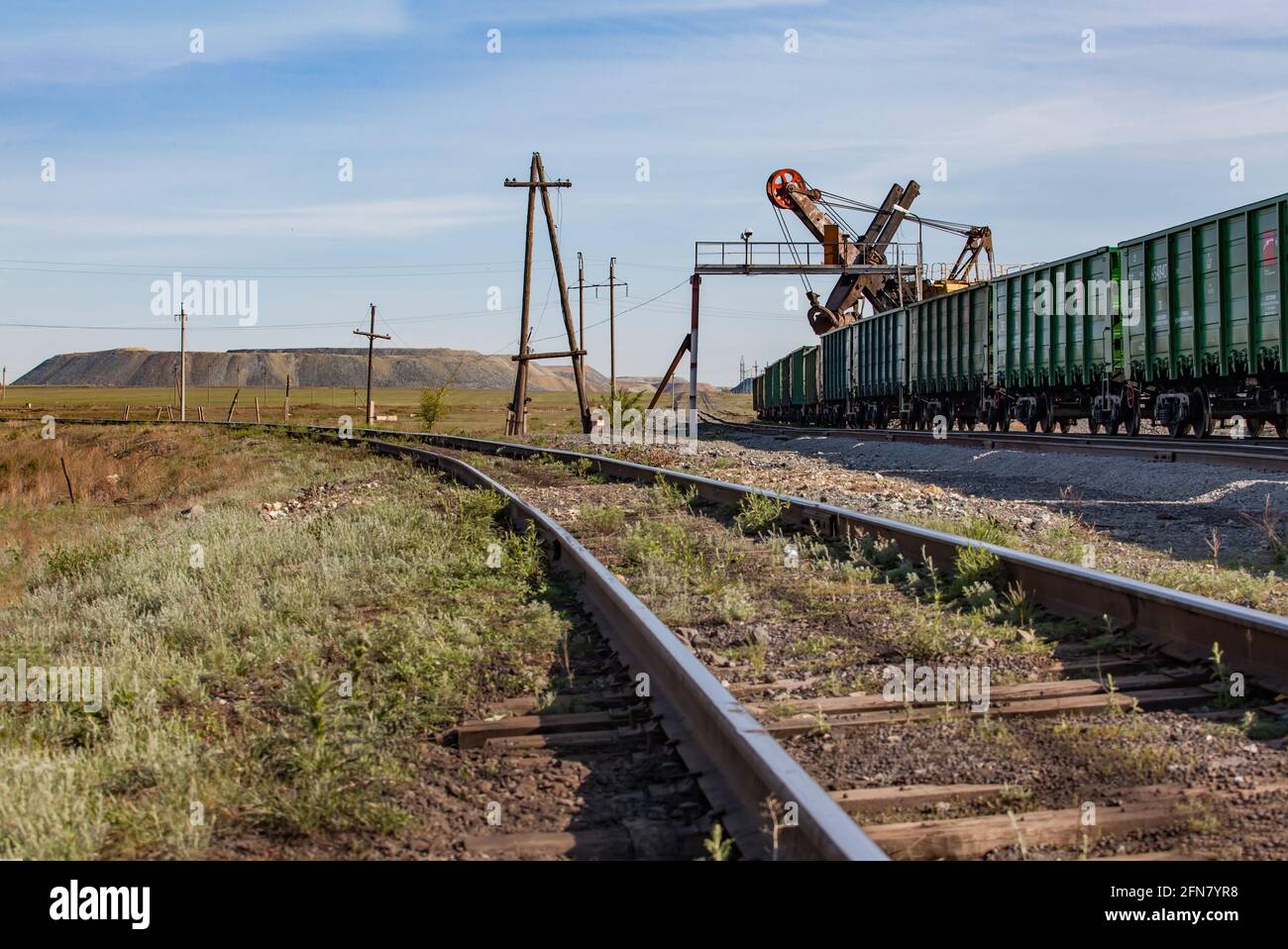 Khromtau, Kasachstan: Erzladestation.Alter Holzmast.Bagger lädt offene Waggons.Wüstensand und Gras.Dump Felsen links am Horizont.Blauer Himmel Stockfoto