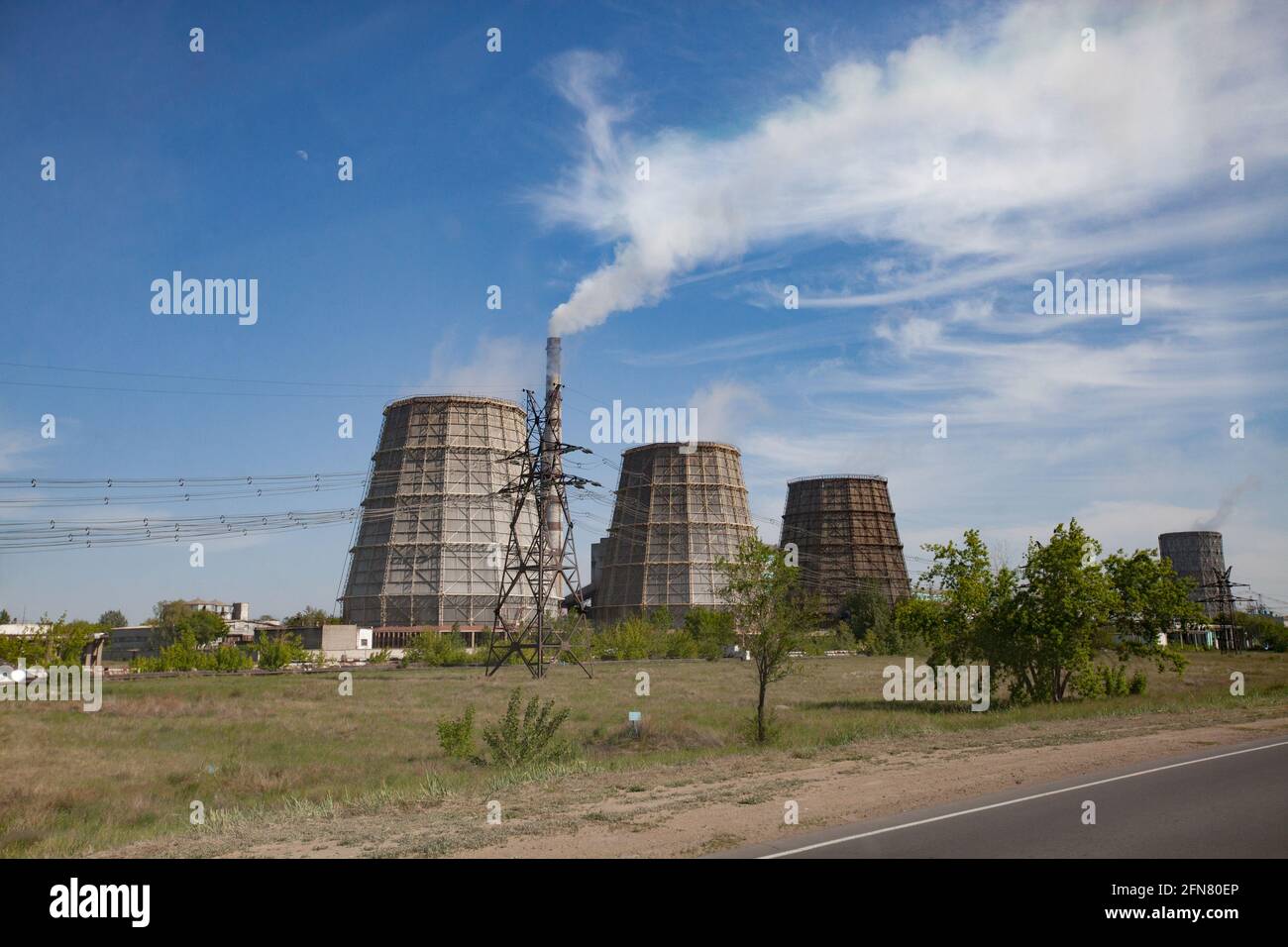 Pavlodar-Wärmekraftwerk. Kühltürme und Rauchschwaden mit weißem Rauch. Blauer Himmel. Grünes Gras, Bäume. Asphaltstraße rechts. Stockfoto