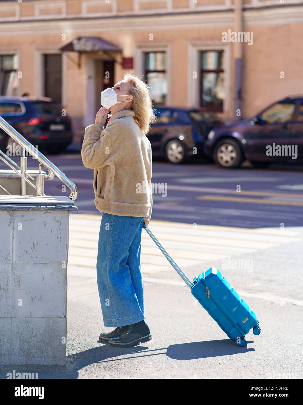 Modische ältere Frau, die in einem Atemschutzgerät oder einer Gesichtsmaske auf einer sonnigen Straße mit blauem Koffer steht und nach oben schaut. Ältere Dame verirrt sich während der Reise während des covid19-Ausbruchs in der Stadt Stockfoto