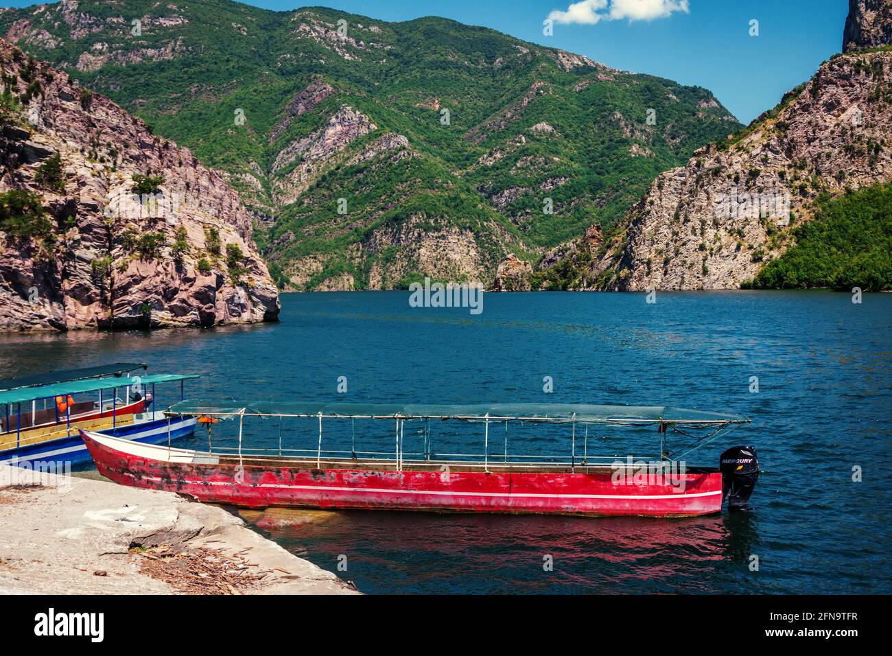 Sommerlandschaft – albanische Berge, bedeckt mit üppigem Laub, See mit ruhigem blauem Wasser und altem roten Boot. Stockfoto