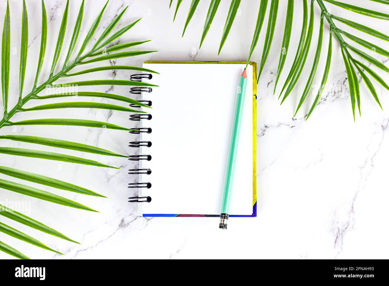 Draufsicht auf flaches weißes Notizbuch mit grünem Bleistift und tropischen Palmblättern auf weißem Hintergrund mit Platz für Kopien. Mock-up und blank temp Stockfoto