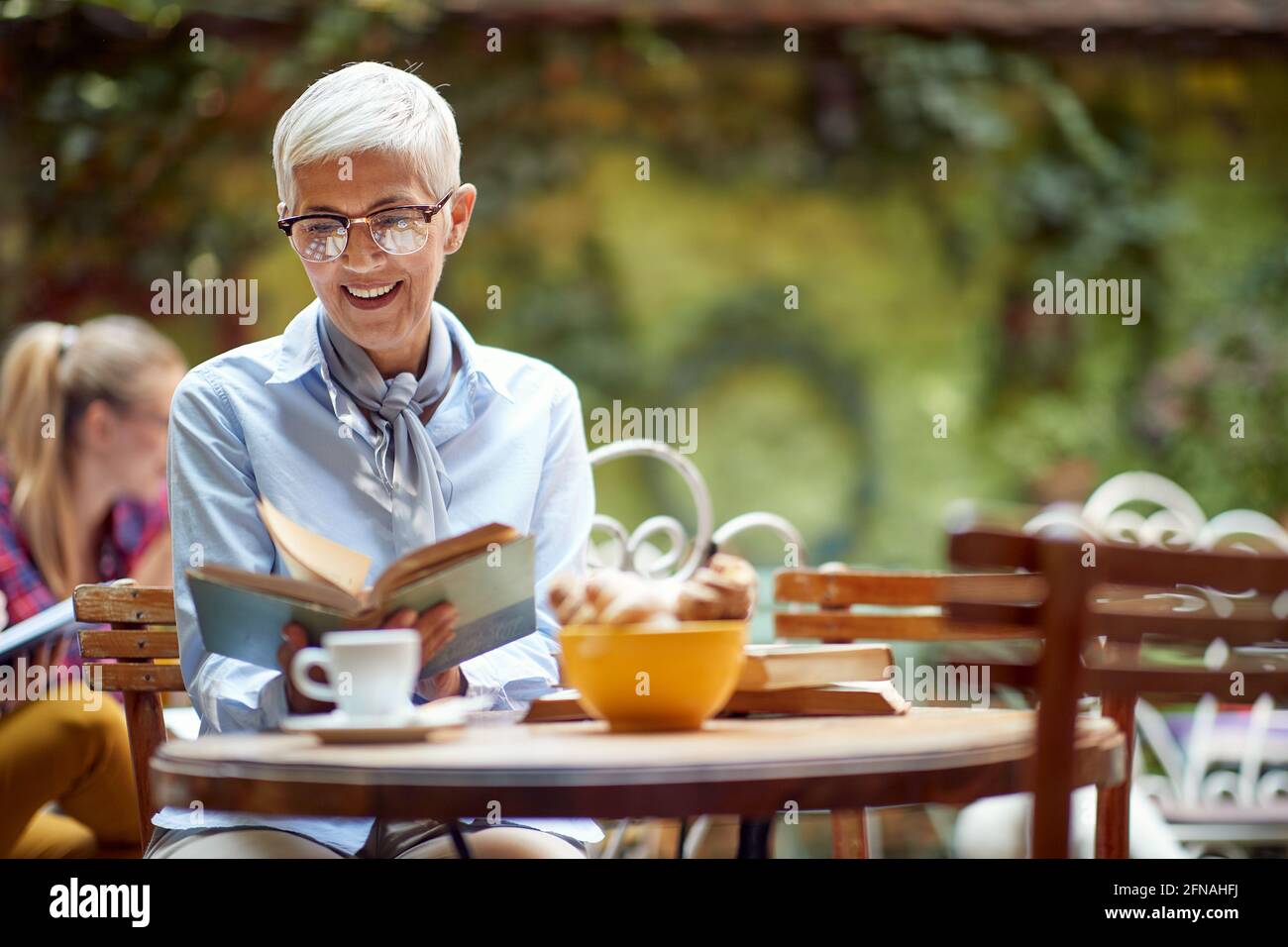 Eine ältere Frau liest ein Buch, während sie einen Kaffee in einer entspannten Atmosphäre in der Bar hat. Freizeit, Bar, im Freien Stockfoto