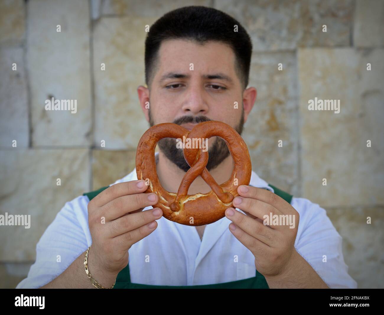 Der junge, ausgebildete kaukasische Bäckermeister schaut sich sein Produkt an, eine traditionelle Laugenbretzel, die Form und Qualität der Laugenbretzel. Stockfoto