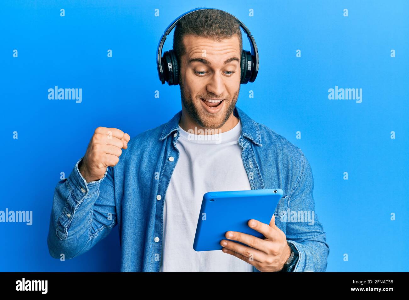 Der junge Kaukasusmann, der mit einem Touchpad und Kopfhörern unterwegs ist, schreit stolz und feiert mit erhobenen Armen Sieg und Erfolg Stockfoto