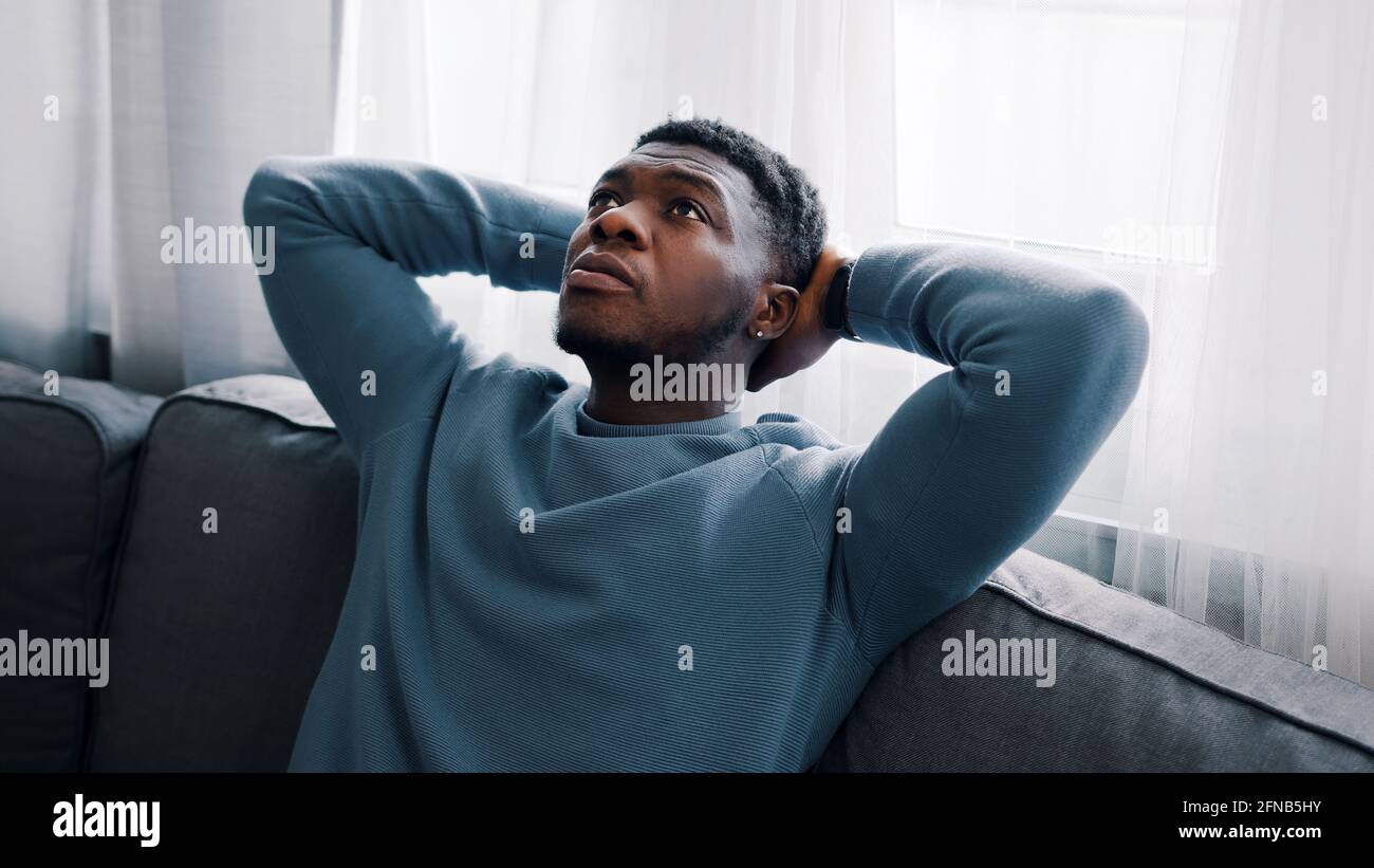 Ernsthafter Mann, der denkt, während er auf einem blauen Sofa sitzt, Hände hinter dem Kopf hält und auf die Decke des Zimmers blickt. Konzentriert. Gemütliches Zimmer mit weißen Vorhängen. Hochwertige Fotos Stockfoto