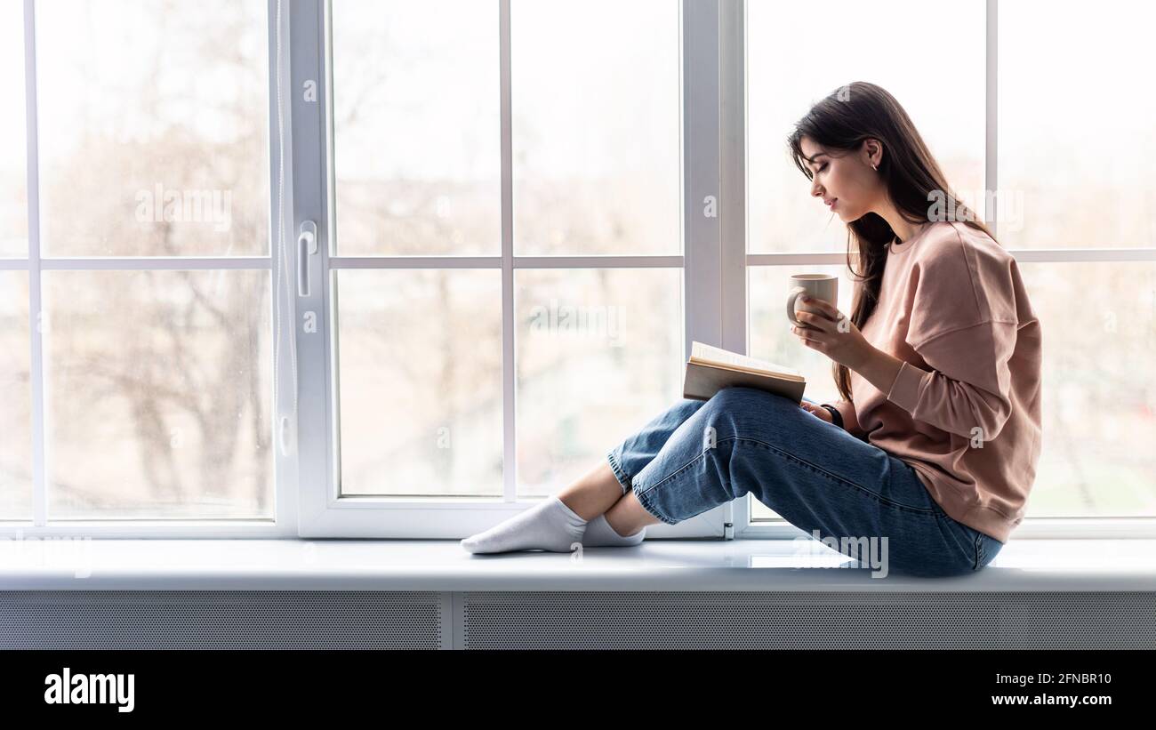 Frau, die auf dem Fensterbrett sitzt und Kaffee trinkt, liest das Buch Stockfoto