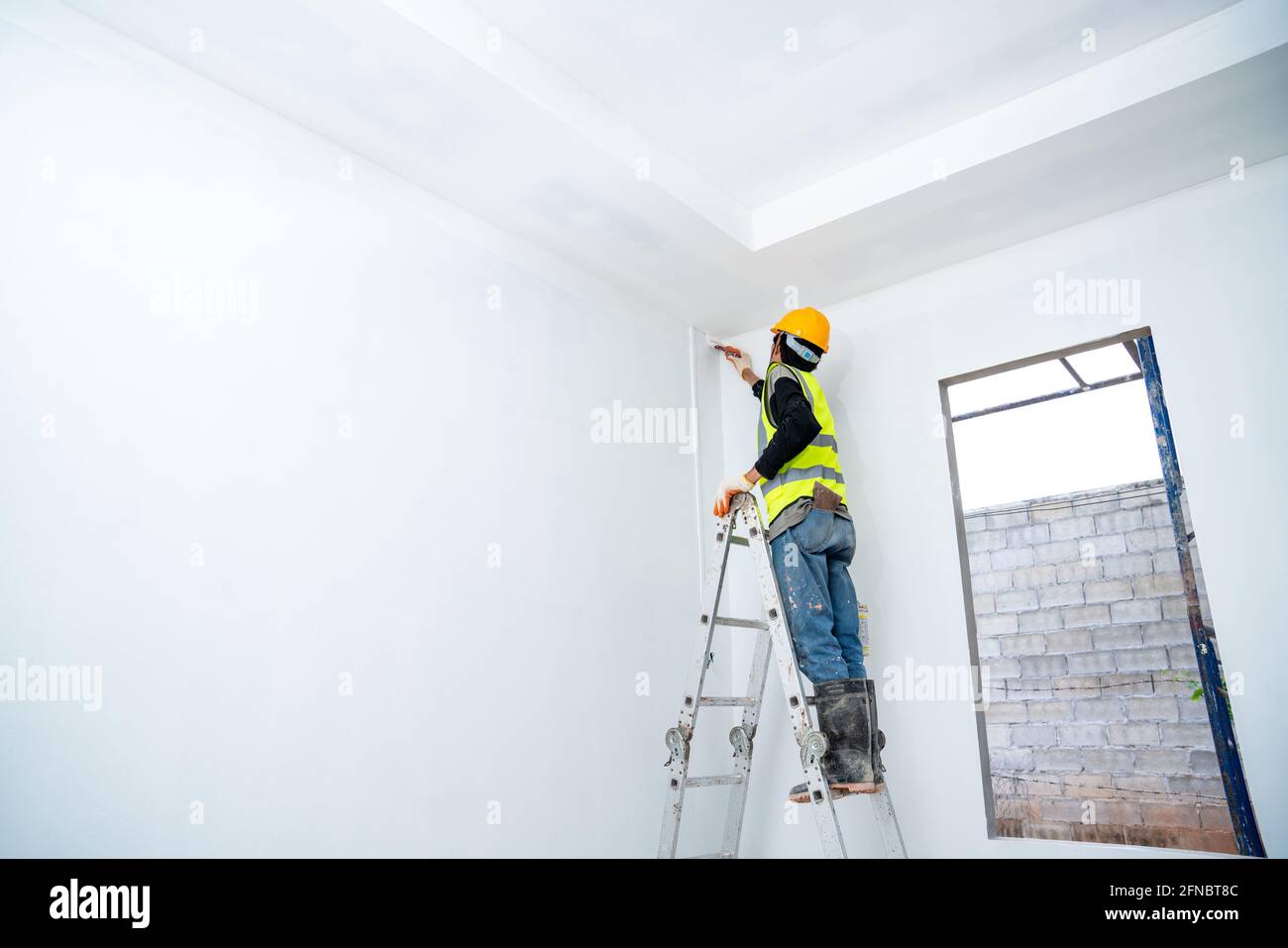 Malermann malte die Wand in einem unfertigen Gebäude, mit Pinsel und Eimer, isoliert auf einem großen leeren Raum mit Leiter auf der Baustelle Stockfoto