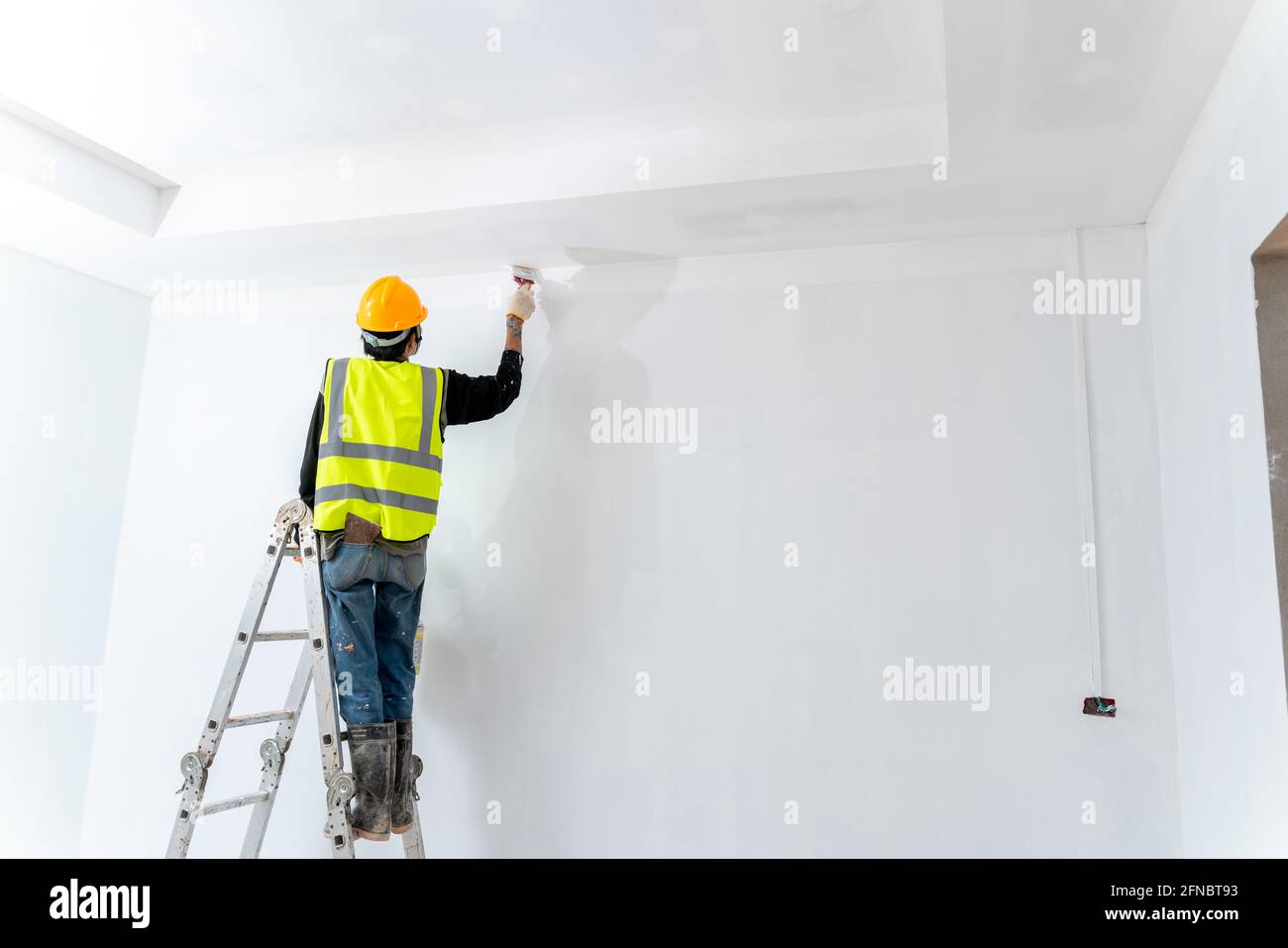 der asiatische Maler Mann malte die Wand in einem unfertigen Gebäude, mit Pinsel und Eimer, isoliert auf einem großen leeren Raum mit Leiter an der constructio Stockfoto