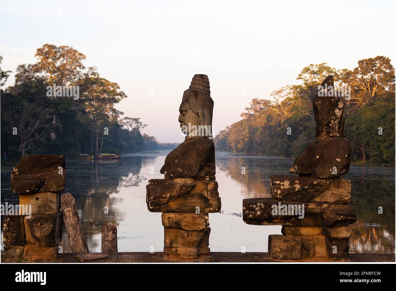 Nahaufnahme einer der Statuen, die den Damm über dem Graben zum Südtor von Angkor Thom säumen. Der Graben ist im frühen Morgenlicht flach ruhig Stockfoto