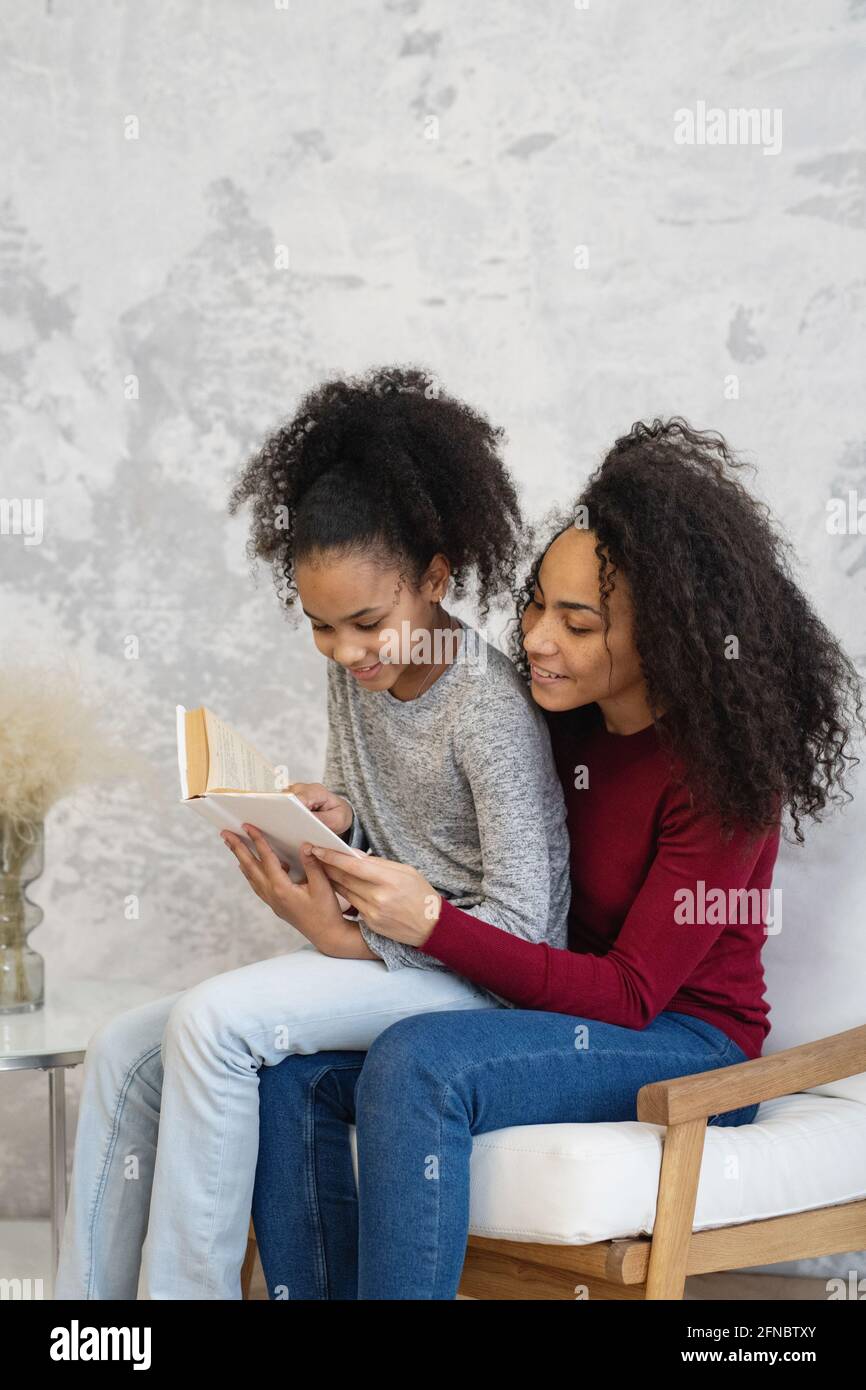 Afroamerikanische Mutter mit ihrer Tochter, die Buch liest, sitzt zu Hause auf einem Stuhl. Stockfoto
