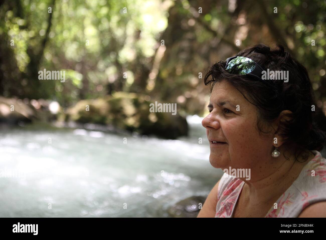 Profil einer lächelnden 62-jährigen älteren Frau im Wald mit dem plätschernden Fluss Banias, einer der drei Quellen des Jordan. Stockfoto