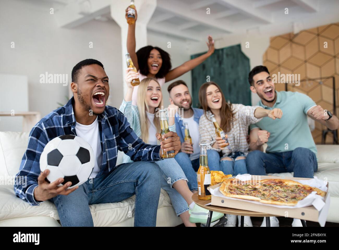 Emotionale multirassische Freunde, die zu Hause Fußball spielen, das Tor schreiend, den Sieg feiern Stockfoto