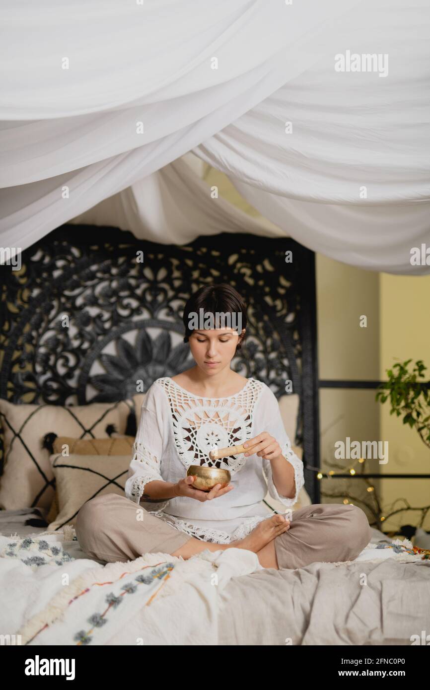 Junge Frau mit tibetischer Klangschale und Stößel auf dem Bett sitzend. Stockfoto