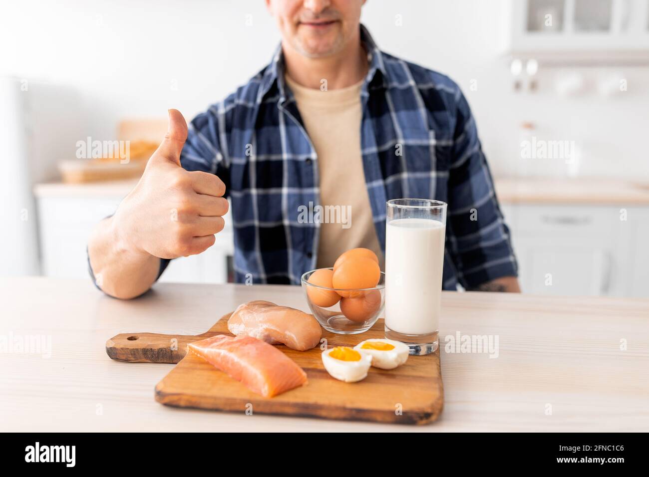 Ich mag gesunde Ernährung. Älterer Mann, der das Frühstück vorbereitet und Daumen, Milch, rohes Fleischfilet, Fisch und Eier zeigt Stockfoto
