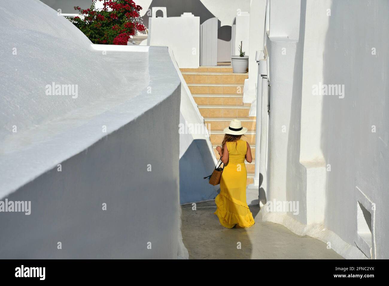 Ein junges Mädchen in einem leuchtend gelben Kleid und einem Panamahut, das auf einer schmalen, weiß getünchten Gasse des Dorfes Oia auf der Insel Santorini, den Kykladen, Griechenland, spaziert Stockfoto