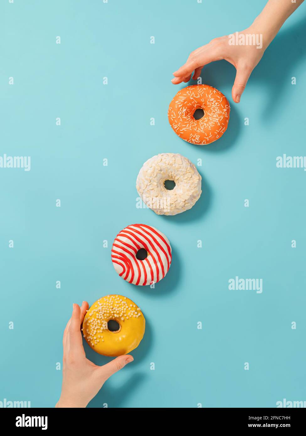 Zwei Hände greifen nach Donuts auf blauem Hintergrund, Draufsicht. Kreatives Layout mit köstlichen glasierten Donuts. Vertikale flache Lay mit Diagonale aus Donuts oder Donuts Stockfoto