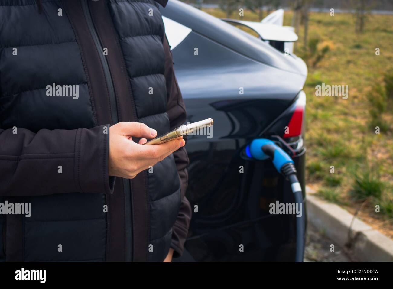 Mann überprüft sein Mobiltelefon, während sein Elektroauto aufgeladen wird. Schnelles EV-Kfz-Ladegerät. Warten auf das Aufladen des EV-Fahrzeugs. Laden von Elektrofahrzeugen. Soziale Medien überprüfen Stockfoto