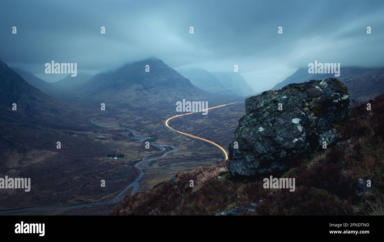 Morgenlandschaft mit Autolampen auf der Bergstraße, die nach Glencoe führt, und einem großen Felsen im Vordergrund. A82 Straße gegenüber Buachaille Etive Mor vor Sonnenaufgang. Schottland Stockfoto