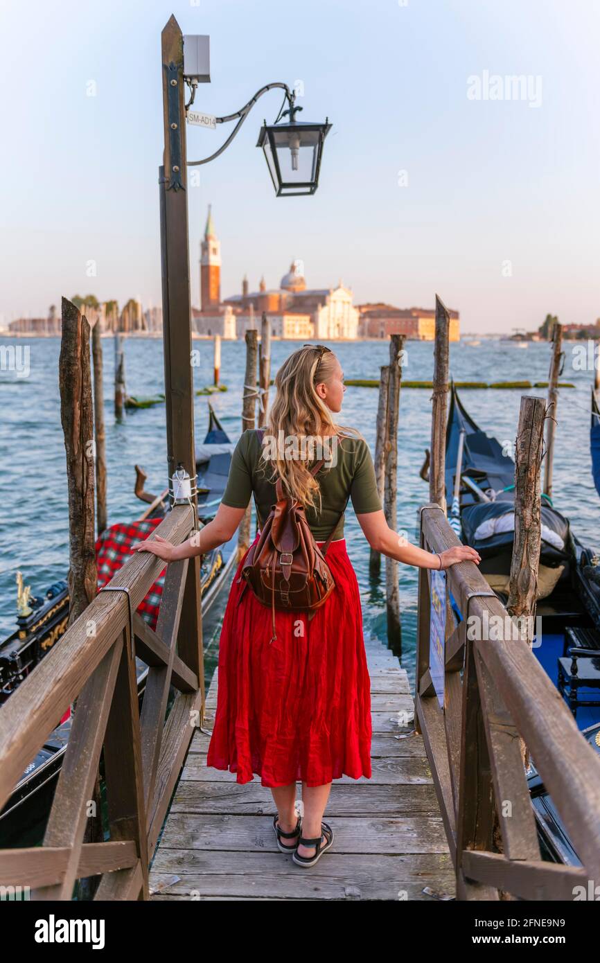 Junge Frau mit rotem Kleid auf einem Steg, venezianische Gondeln, in der hinteren Kirche San Giorgio Maggiore, Venedig, Venetien, Italien Stockfoto