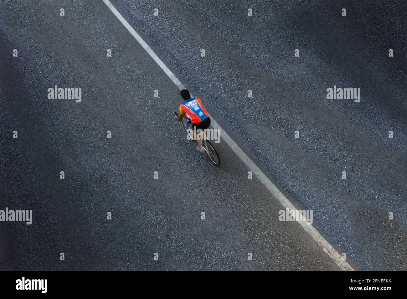 Einsam verschwommener Radfahrer oder Biker mit Hemd, aber ohne Helm auf einer Asphaltstraße mit nur einer weißen Linie, die diagonal im Foto ist. Blick von oben Stockfoto