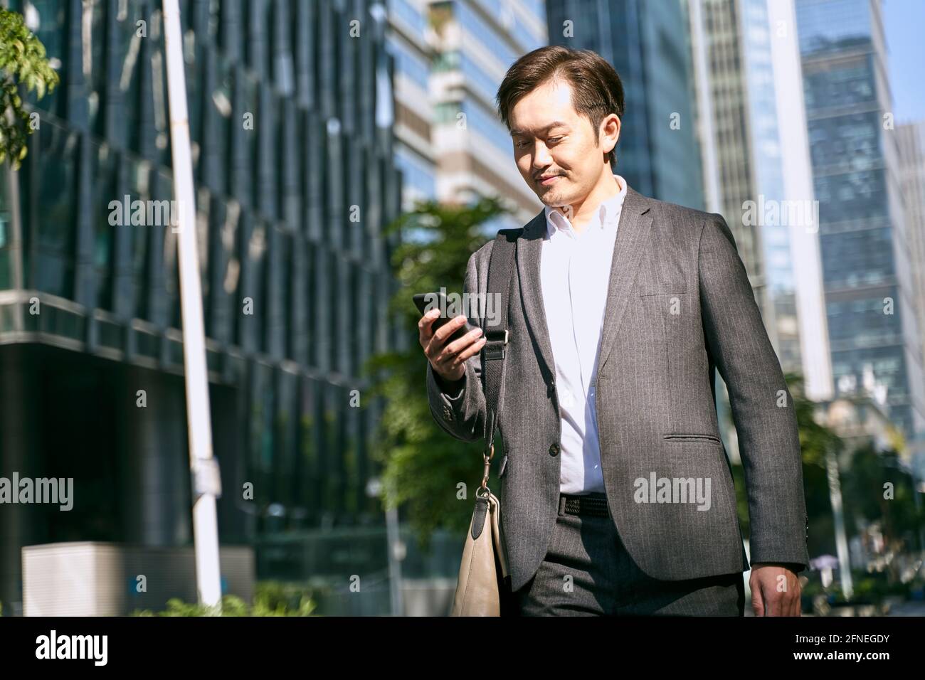 asiatischer Büroangestellter mit weißem Kragen, der während des Telefonanschlosses auf das Mobiltelefon schaut Zu Fuß zum Finanzviertel der modernen Stadt zu arbeiten Stockfoto