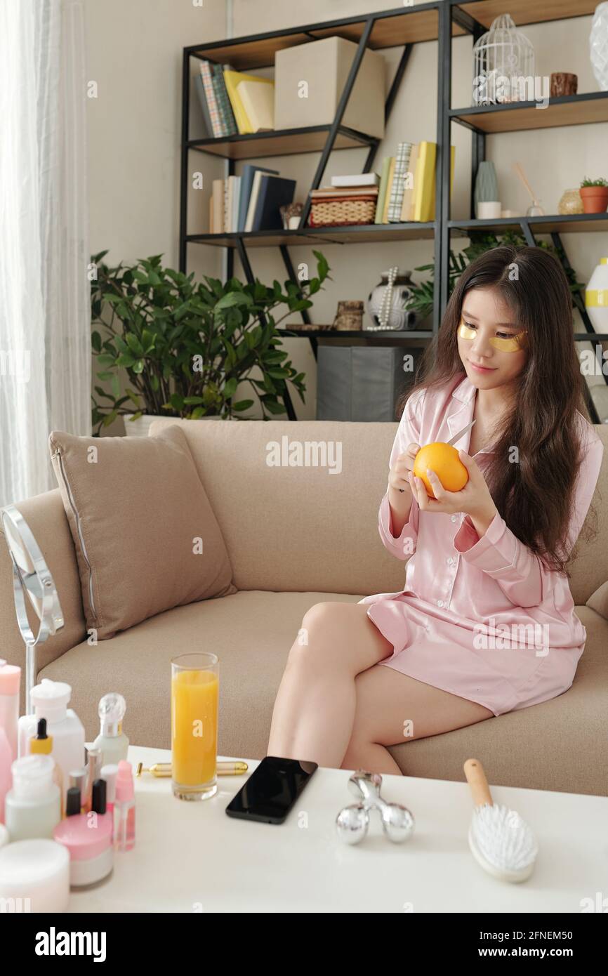 Hübsche junge Frau im Schlafanzug sitzt auf dem Sofa mit Unterauge Aufnäher und schneiden von Orange zum Frühstück Stockfoto