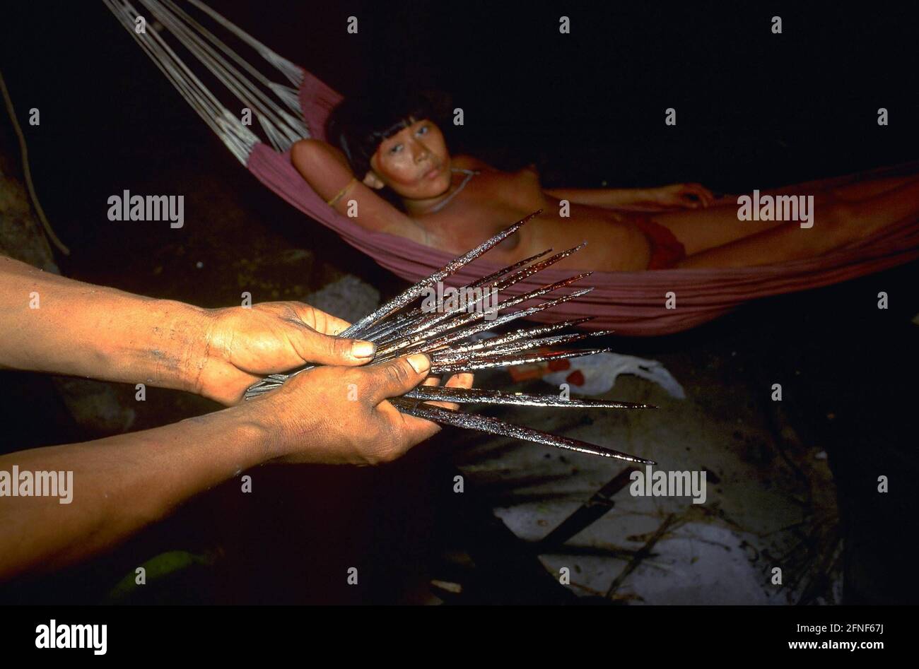 Der Mann des Yanomami-Stammes hält sogenannte Curare-Pfeile in seinen Händen. Diese Pfeile enthalten ein tödliches Gift. Im Hintergrund eine Frau in einer Hängematte. [Automatisierte Übersetzung]' Stockfoto