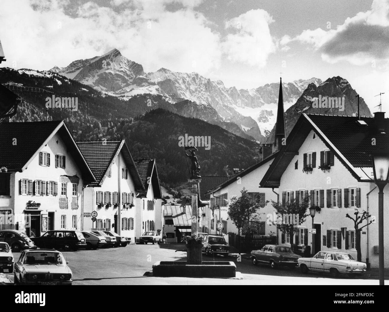 'Dieses undatierte Foto, aufgenommen um 1975, zeigt den Floriansplatz mit dem Floriansbrunnen in Partenkirchen, der Blick geht in die Ballengasse. An einer Ampel rechts hängt ein Schild mit der Aufschrift: ''zum öffentlichen WC 150 m''. Auf der linken Seite befindet sich ein Friseursalon. Rechts erhebt sich hinter den Häusern der Turm der Kirche Mariä Himmelfahrt. Im Hintergrund ragt das Wettersteingebirge mit dem Rießerkopf (vorne, Mitte des Bildes) und den schneebedeckten Gipfeln der Alpspitze, Höllentalspitzen, Zugspitze und Waxenstein (Hintergrund, Stockfoto