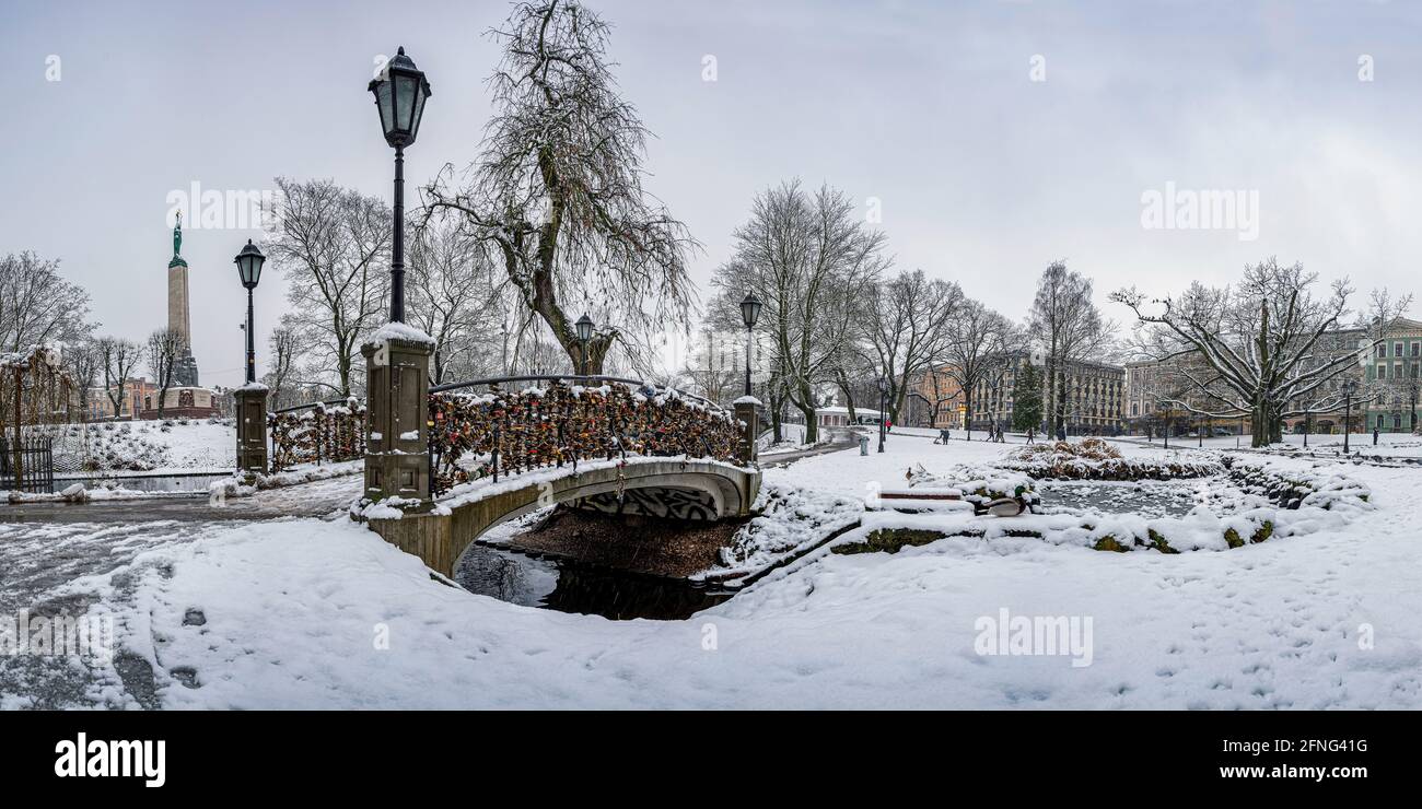 Blick auf den Bastion Hill Park und das Freiheitsdenkmal in Riga, Lettland. Winterlandschaft in verschneiten Park mit schöner kleiner Brücke mit Schleusen über Teich. Stockfoto