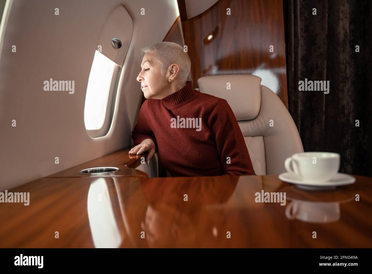 Reife Frau, die das Fenster des Privatjets fast verschwommen betrachtet tasse Kaffee Stockfoto