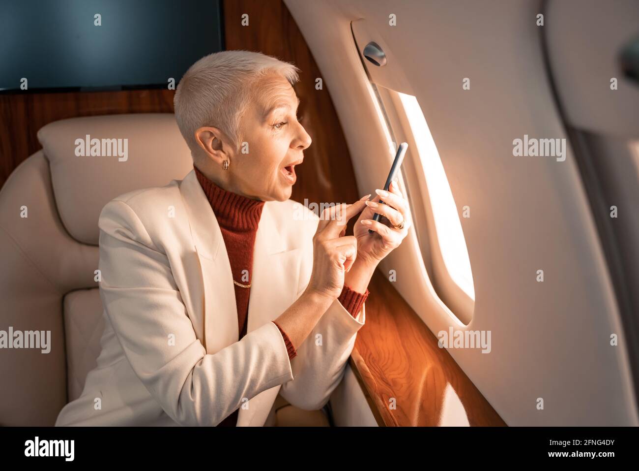 Staunende Geschäftsfrau mit Smartphone, die im Privatflugzeug auf das Fenster schaut Stockfoto