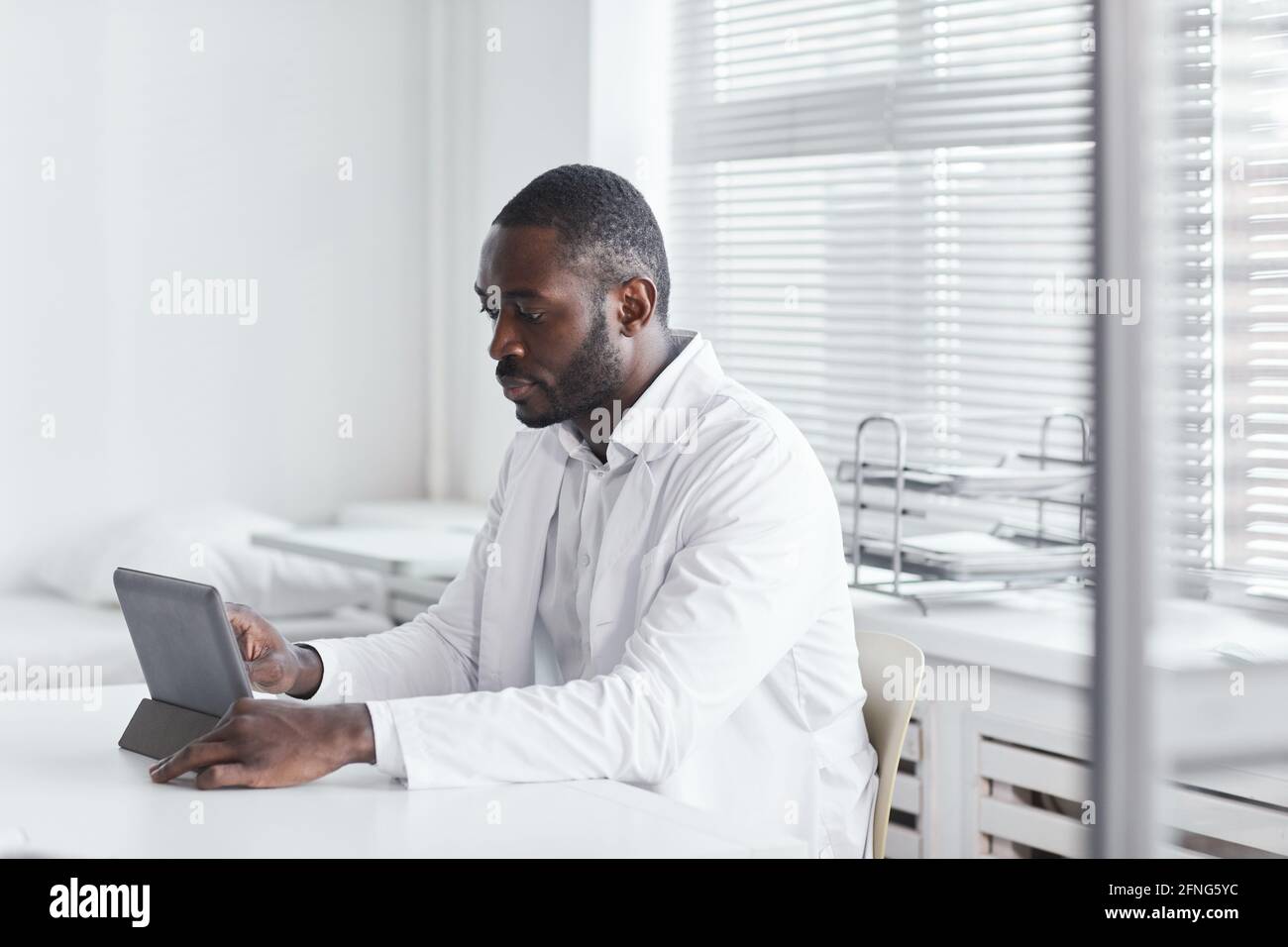 Afrikanischer junger Arzt sitzt am Tisch und arbeitet daran Digitales Tablet in seinem Büro Stockfoto