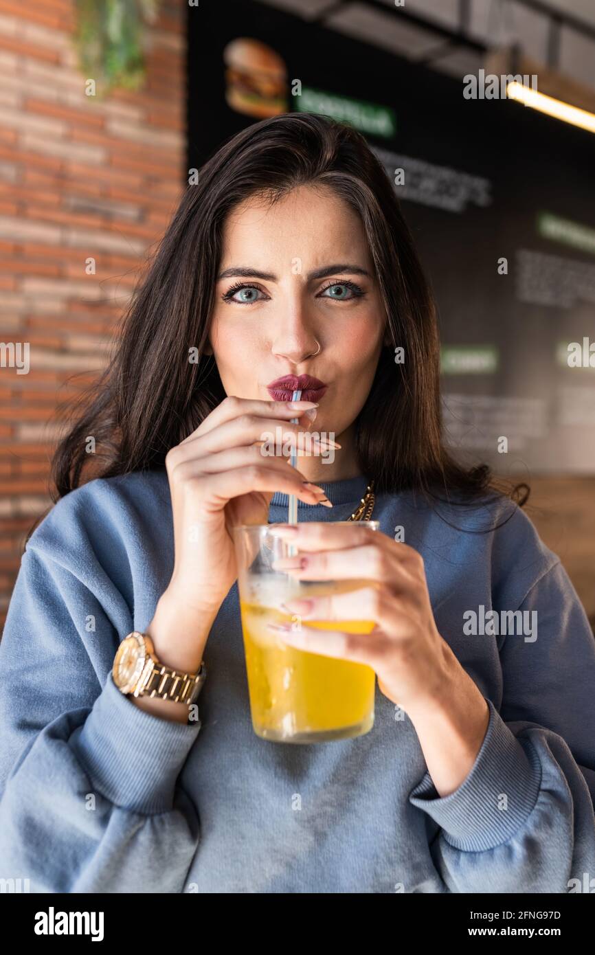 Fröhliche junge Frau in blauen Pullover nippen kalt fizzy weich Trinken Sie durch Stroh, während Sie Zeit in der Cafeteria und verbringen Glücklich auf die Kamera schauen Stockfoto