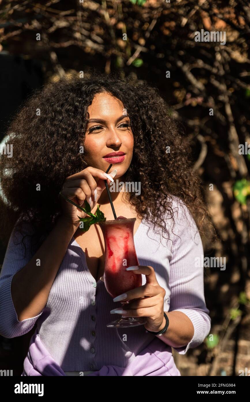 Positive junge ethnische Frauen tragen lässiges Outfit und genießen frische Süße Cocktail und Blick weg, während Sie im sonnigen Garten stehen Stockfoto