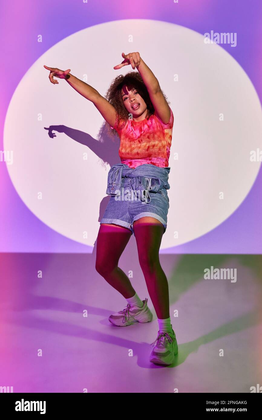 Full Body Fit afroamerikanische Tänzerin in Shorts tanzen Die Arme sind angehoben und die Zunge ist nach außen und blickt dabei auf die Kamera Stehen in Neonlicht in Stud Stockfoto