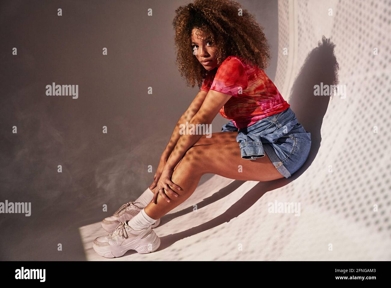 Seitenansicht Vollkörper wunderschöne selbstbewusste afroamerikanische Frau in Shorts sitzen an kurvigen Wänden und blicken auf die Kamera Stockfoto