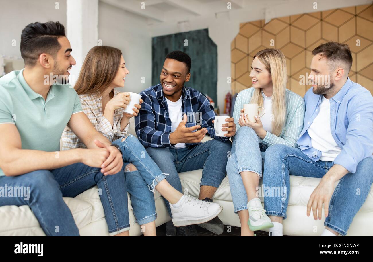 Gruppe von fröhlichen multiethnischen Freunden, die zu Hause feiern, auf der Couch sitzen und Kaffee trinken Stockfoto