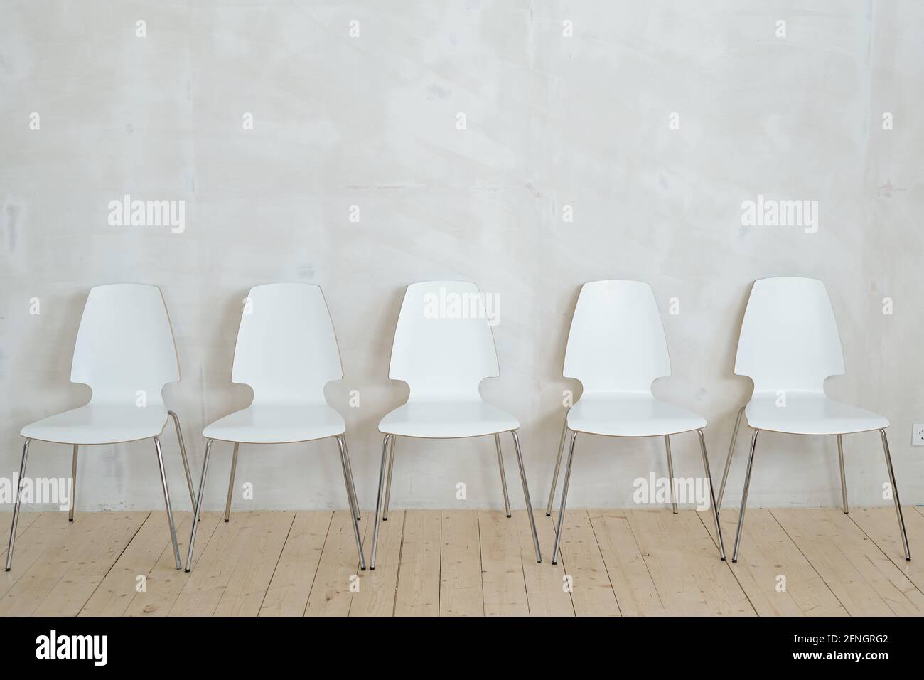 Reihe von weißen Plastikstühlen auf Metallbeinen platziert Parkett gegen graue Wand Stockfoto