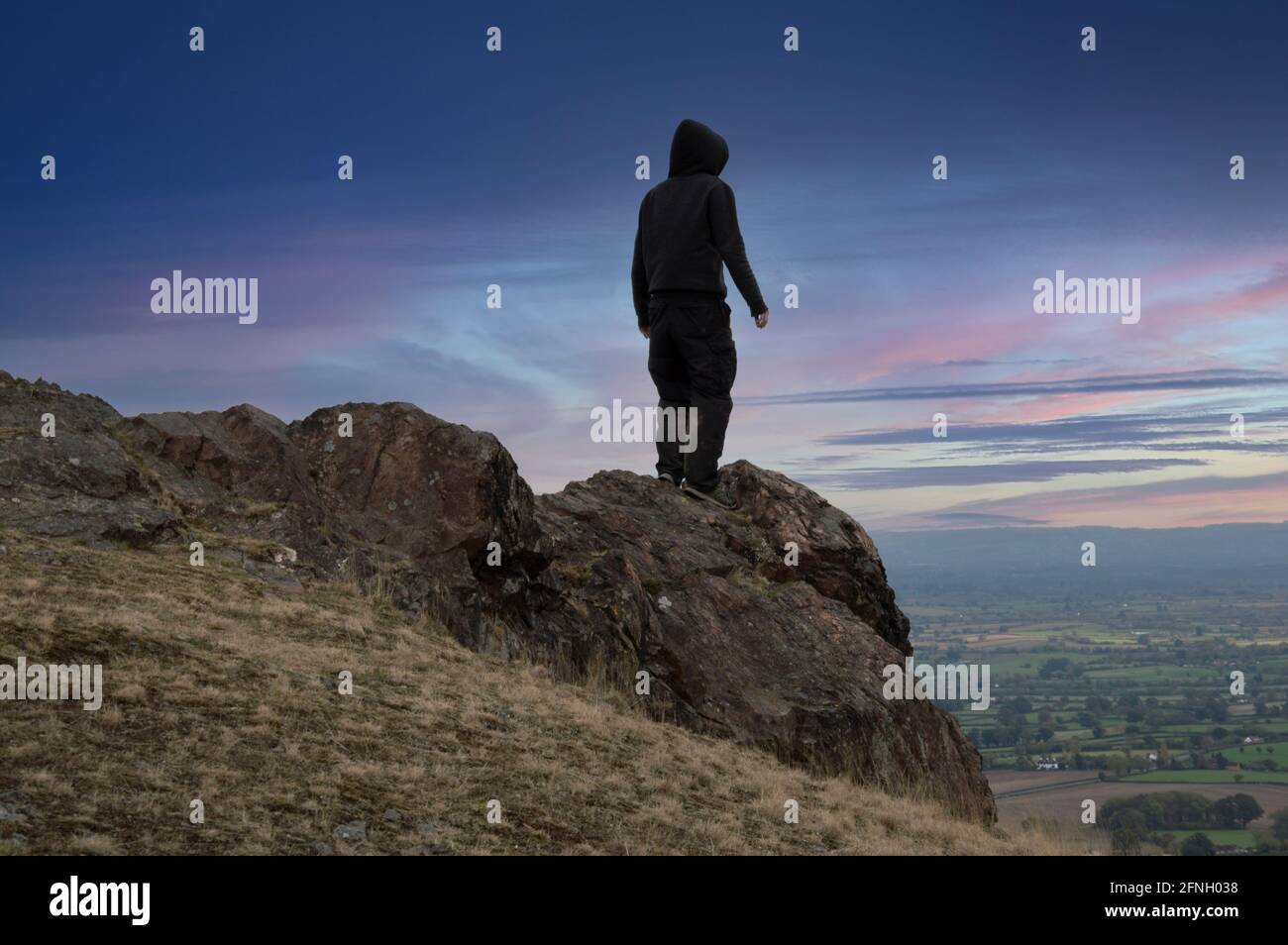 Ein launischer Mann mit Kapuze, zurück zur Kamera, der auf einem Berg steht und einen wunderschönen Sonnenuntergang anschaut. Stockfoto