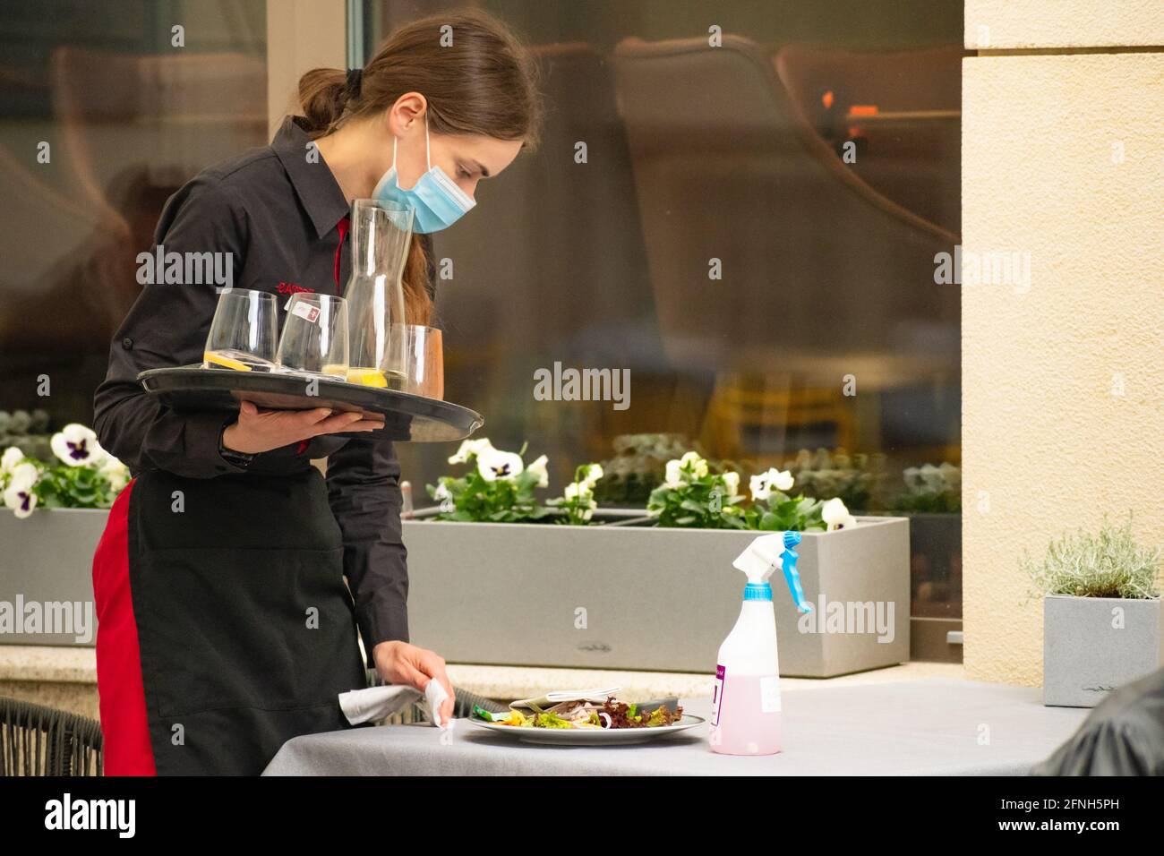 Hübsche Kellnerin mit einer Maske, die den Tisch einer Bar, eines Cafés oder Restaurants im Freien aufräumte, reinigte und desinfizierte und nach der Sperre wieder öffnete Stockfoto