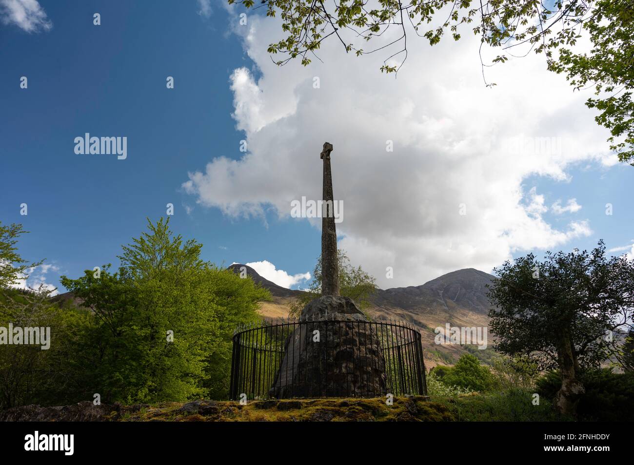 Glencoe Massacre Gedenkkreuz im Dorf Glencoe, Schottische Highlands an einem sonnigen Tag mit blauem Himmel und Wolken. Stockfoto