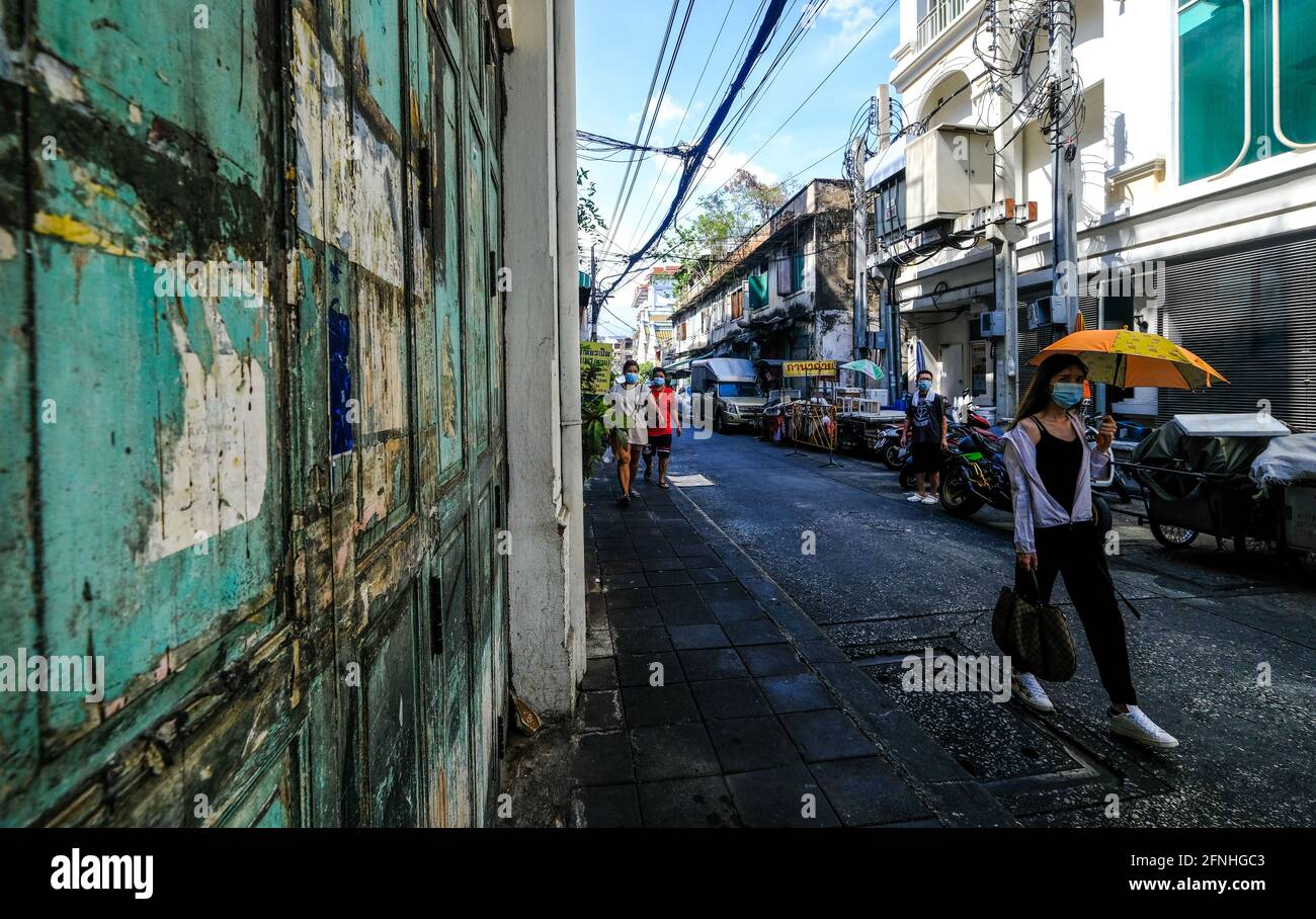 Eine junge Frau, die einen Gesichtsbesen trägt und einen orangefarbenen Regenschirm hält, geht an einer alten, grün verwitterten Tür in der Chinatown-Gegend von Bangkok, Thailand, vorbei Stockfoto