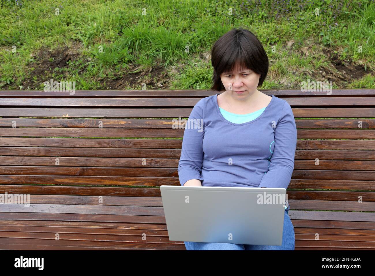 Frau, die mit einem Laptop auf ihrem Schoß auf einer Holzbank im Park sitzt. Konzept für Remote-Arbeiten im Freien, freiberuflicher Mitarbeiter Stockfoto