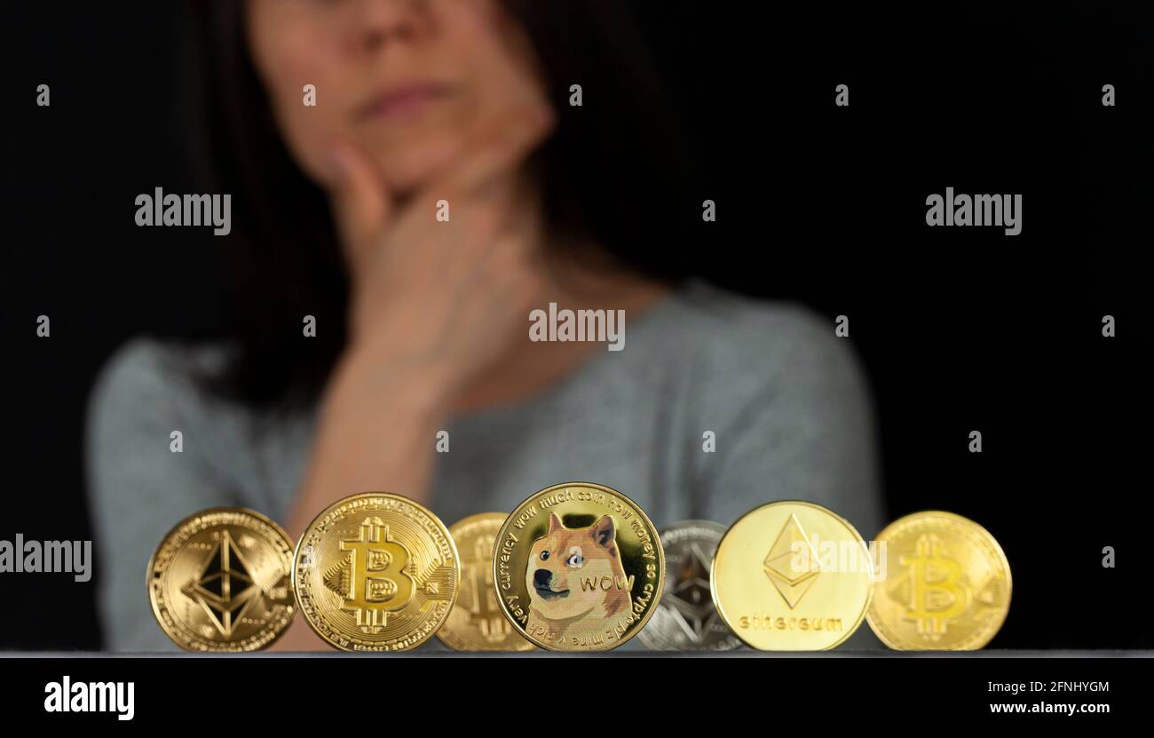 Eine Vielzahl von trendigen symbolischen Kryptowährungstypen, die als glänzende Münzen dargestellt werden, stehen vor dunklem Hintergrund. Eine Frau mit der Hand auf dem Kinn ist un Stockfoto