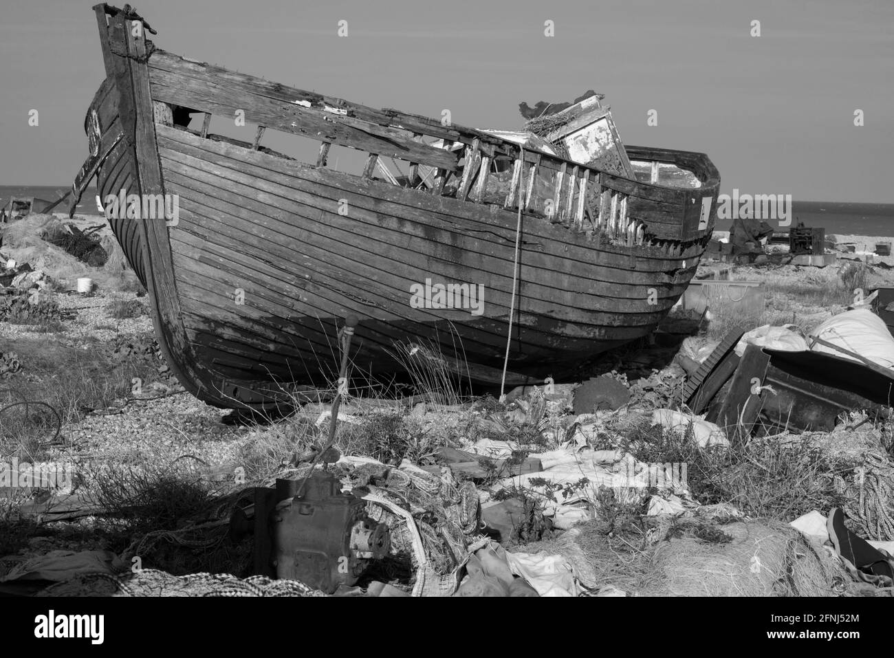 Monobild des kleinen Fischerbootes alt und hoch und trocken und verrottet auf einem Kiesstrand, umgeben von zerbrochenen, rostenden und weggeworfenen Geräten Stockfoto