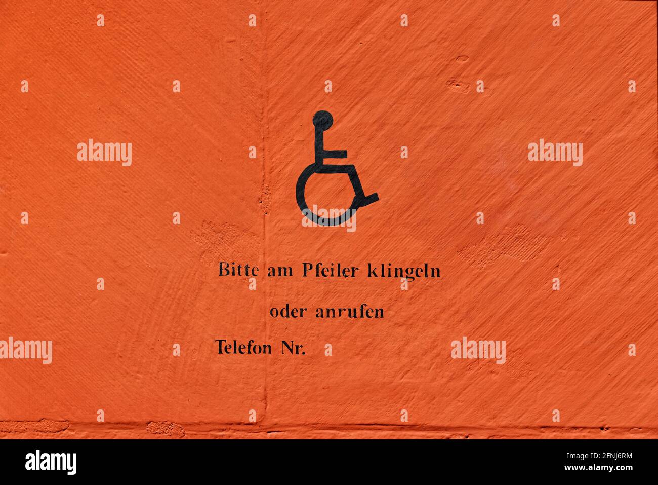 Behinderten-Symbol an einer orangefarbenen Wand mit deutschem Text, deutsche Übersetzung: Bitte klingeln Sie an der Post oder rufen Sie an, Telefonnummer Stockfoto