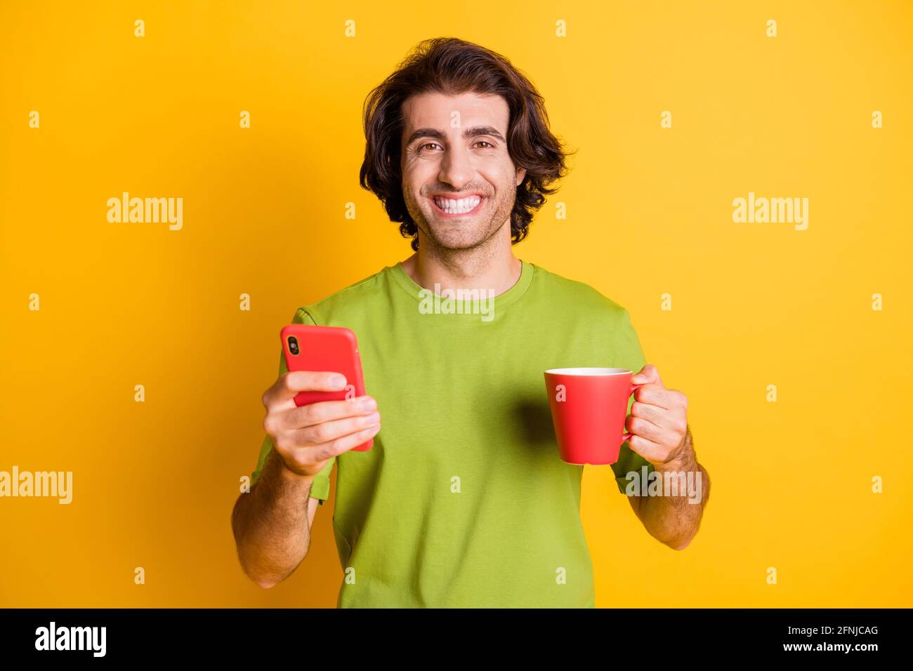 Porträtfoto des Mannes, der mit dem Smartphone die Tasse mit Cappuccino hält Lächelnd isoliert auf lebhaft gelbem Hintergrund Stockfoto